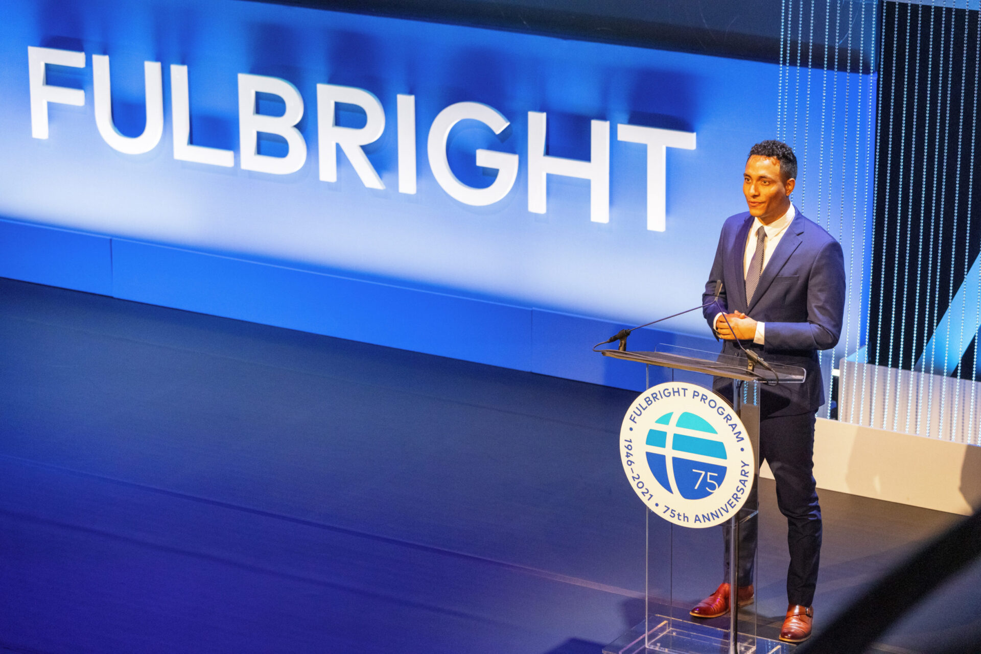 A person standing at a podium in a suit. The podium has a Fulbright logo on it and next to the person is "Fulbright" in big letters on the stage.