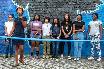 Girls standing in front of wall and woman speaking into microphone behind blue ribbon. 