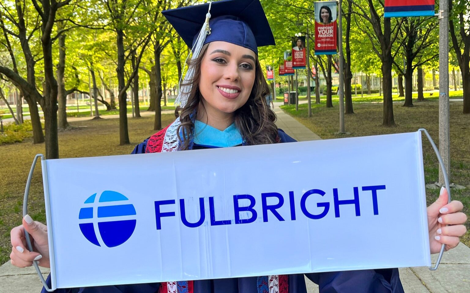 Fulbright alum standing in park at University of Chicago, Illinois in cap and gown, holding Fulbright sign.