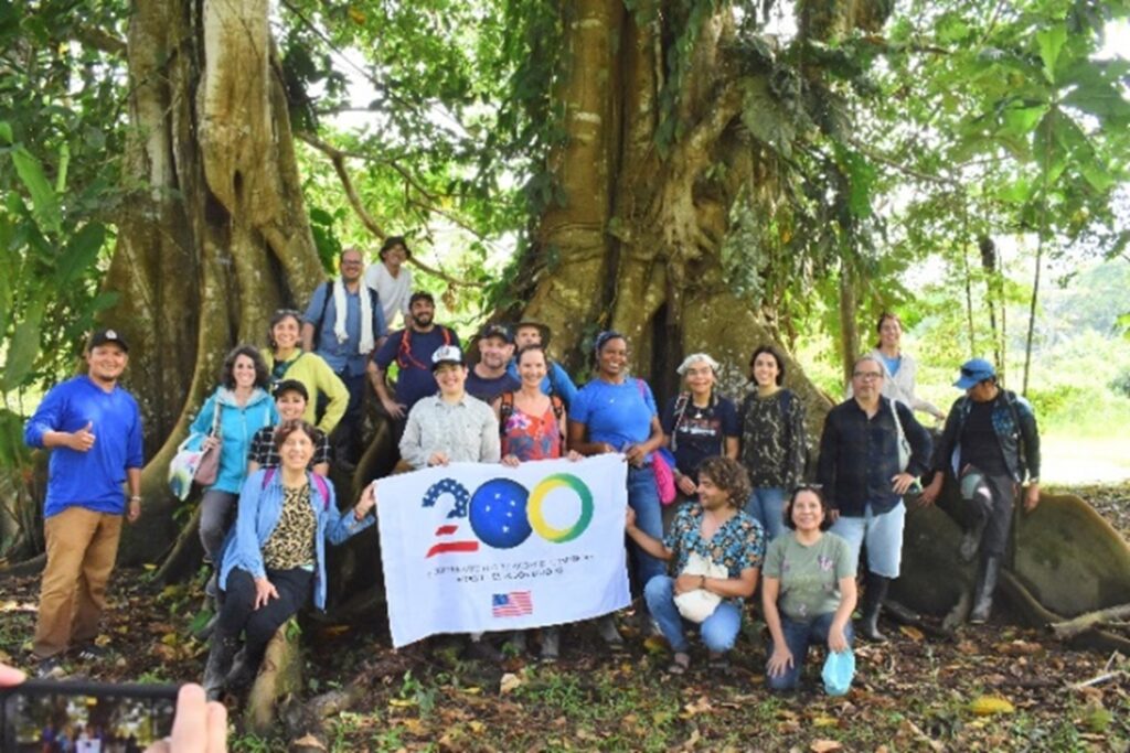 Amazonian Scholars standing under trees with sign celebrating Bicentennial of US–Brazil diplomatic relations.