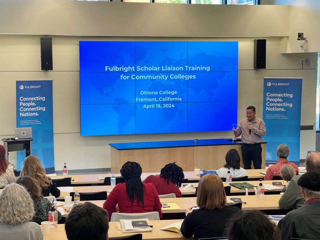 Scholar Liaison, Charles Sasaki, standing in front of projector screen presenting to other scholar liaisons in lecture room.