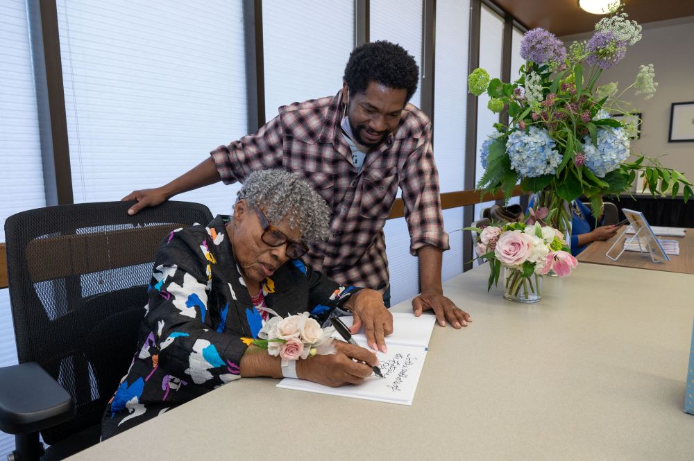Opal Lee sitting writing at table with Sedrick Huackaby standing next to her