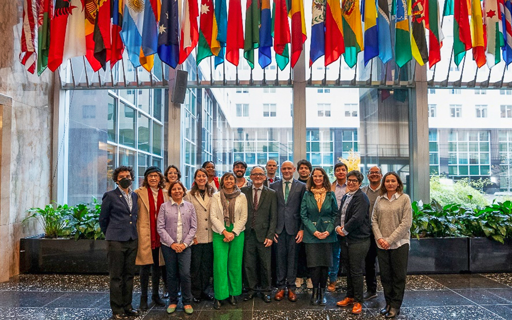 A group of fifteen people standing in front of large windows with international flags hanging above