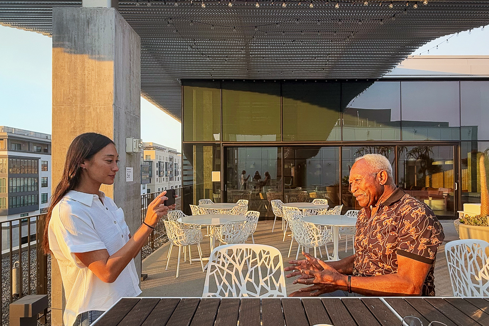 Two people interact on a rooftop patio at sunset, with modern architecture and furniture in the background.