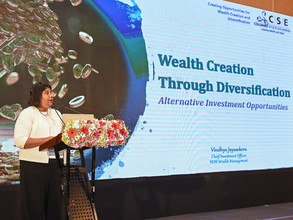 A woman speaks at a podium with a floral arrangement in front of a presentation screen about wealth creation.