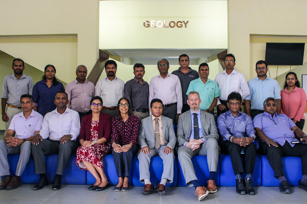 Group portrait of seventeen people in a professional setting below a "GEOLOGY" sign.