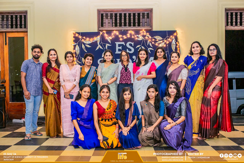 A group of fifteen people posing indoors, wearing colorful attire, with a backdrop of fairy lights and decorative motifs.