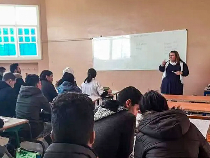 Students in a classroom listening to a teacher near a whiteboard.