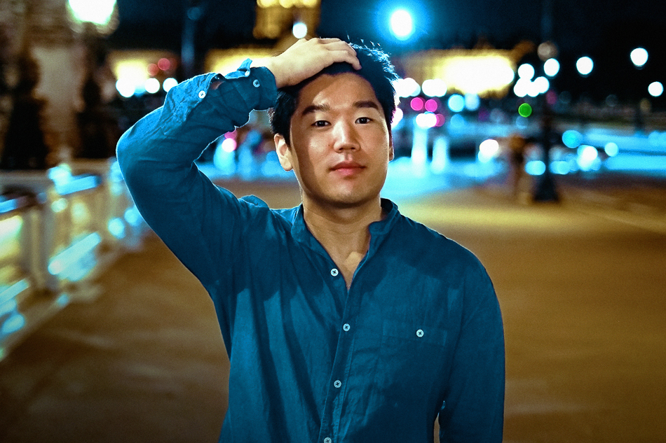 A person in a blue shirt stands outside at night, with colorful lights in the blurred background.