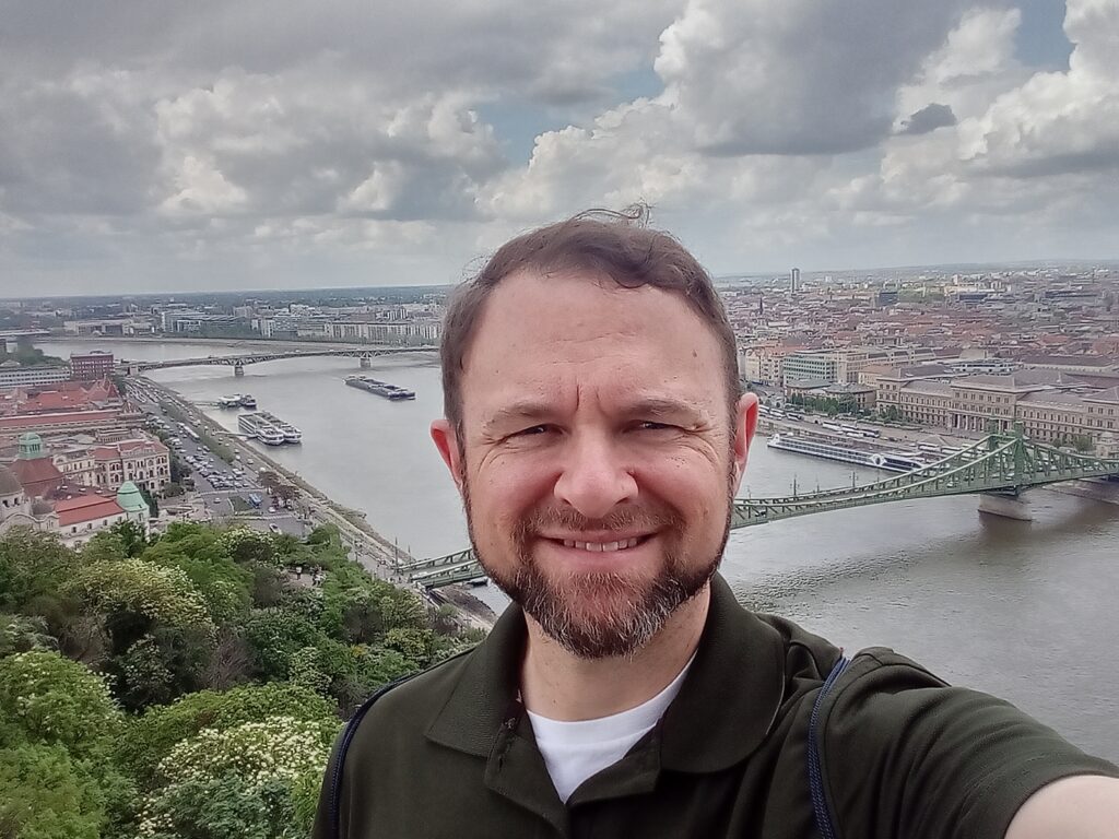 Man smiling with a river, bridge, and cityscape in the background.