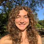 A person with curly hair smiling outdoors with trees and a blue sky in the background.