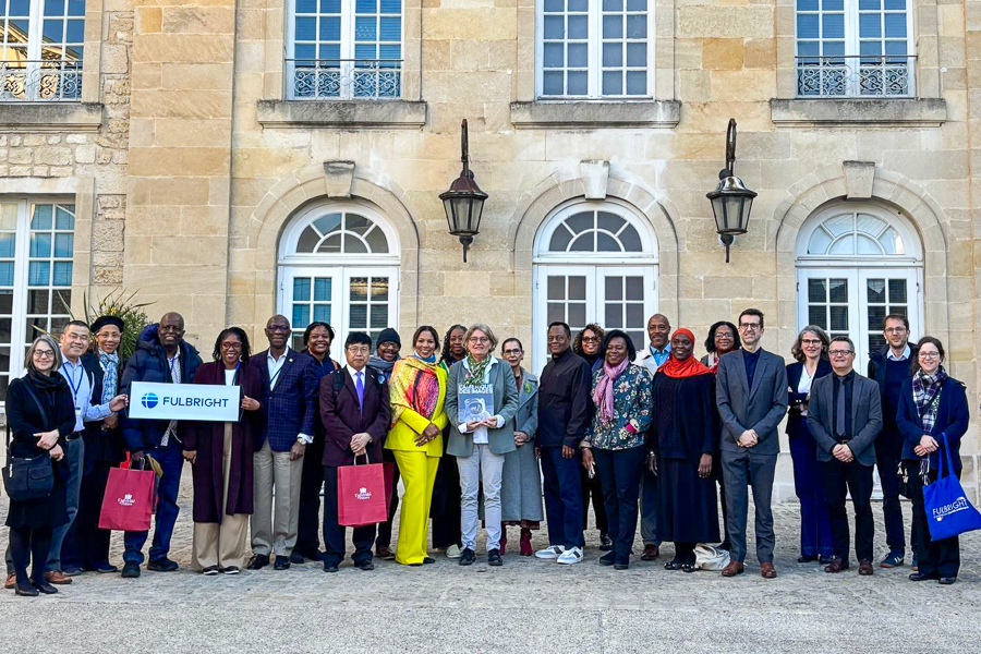 A group of people standing together in front of an arched-windowed stone building, some holding Fulbright signs and bags.