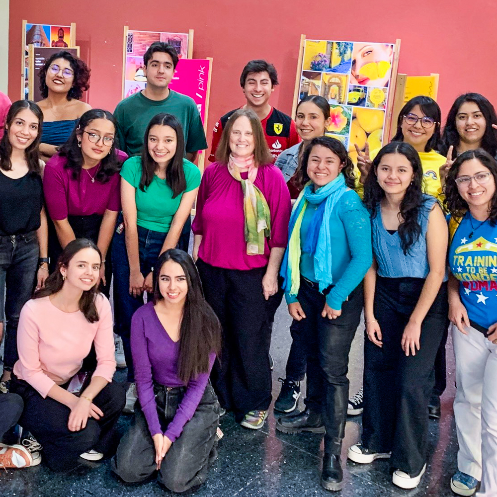 A group of fourteen people posing indoors against a red wall with colorful posters.
