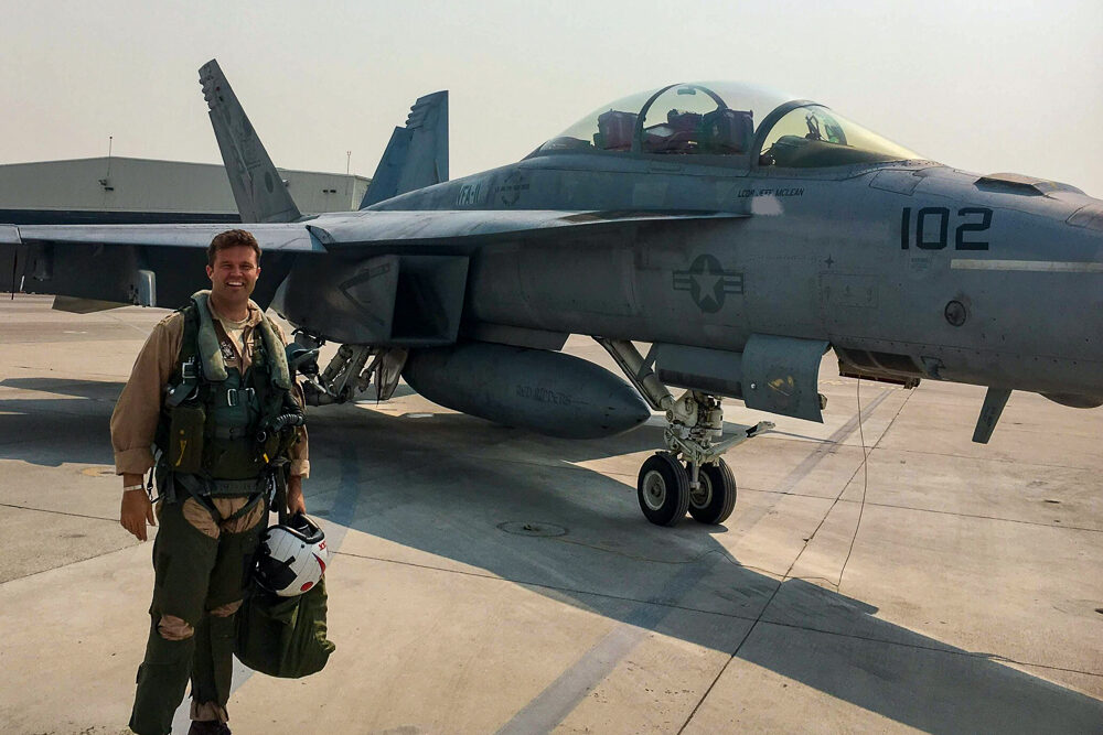 McLean, a pilot stands in front of a gray military fighter jet on a tarmac.