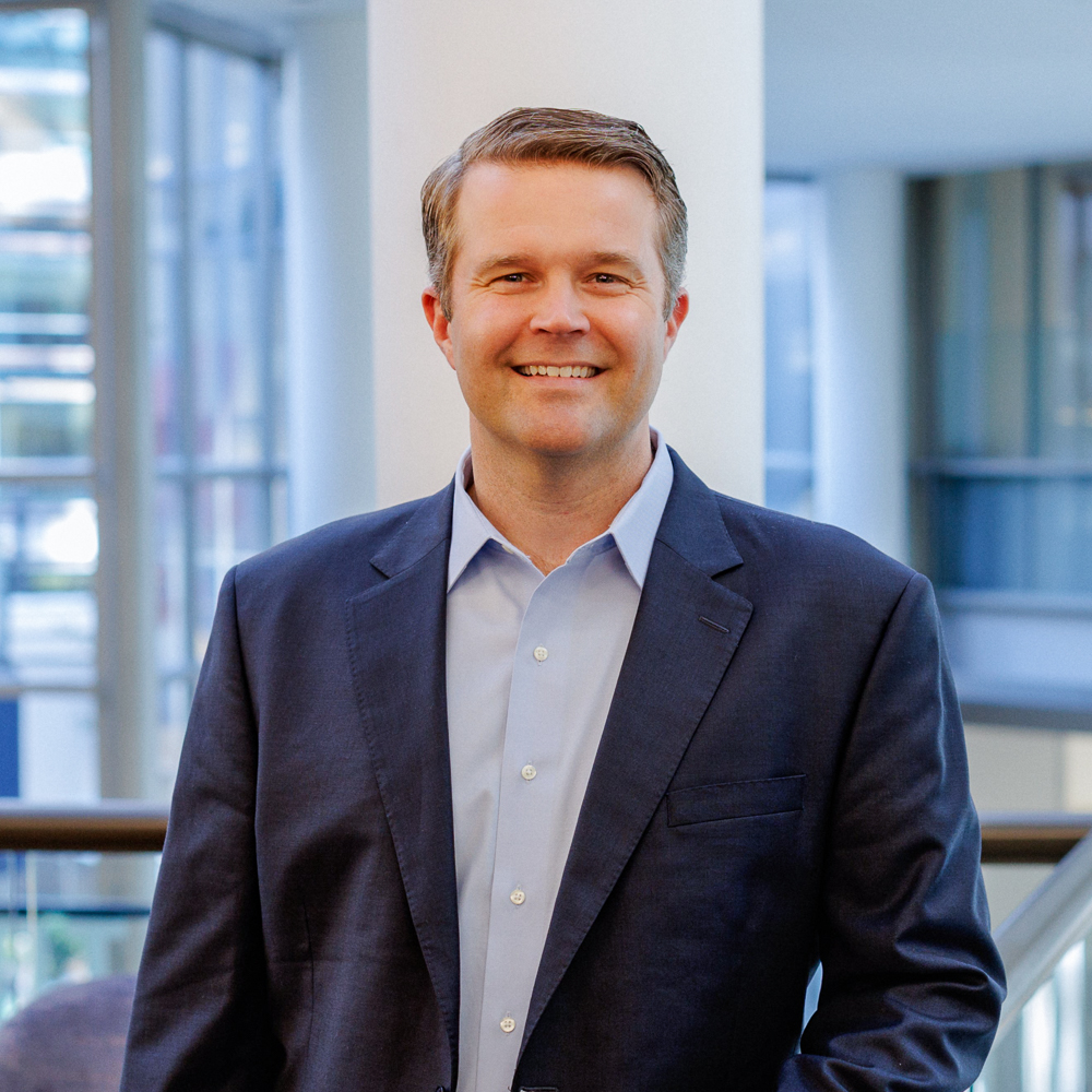 Person in a blue suit smiling indoors with a modern office background.