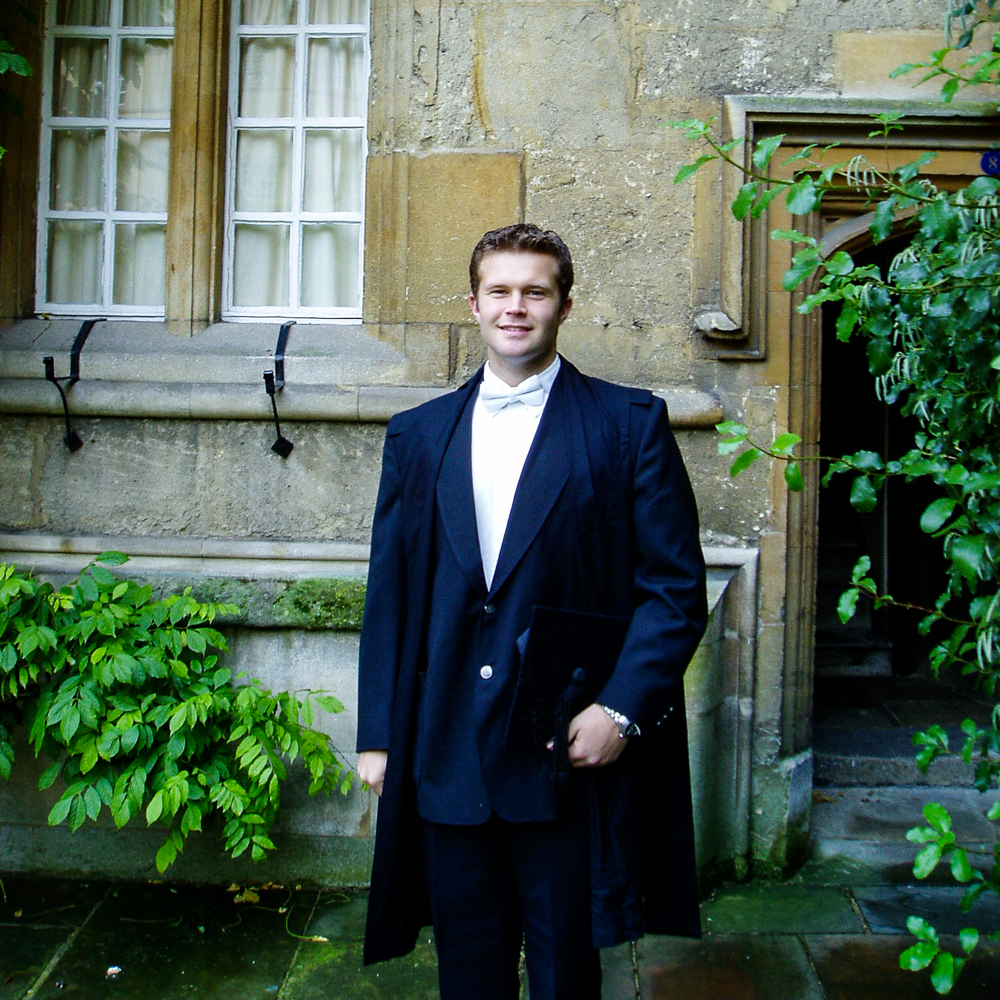 McLean in formal academic attire standing in front of an old stone building with windows and an arched doorway.