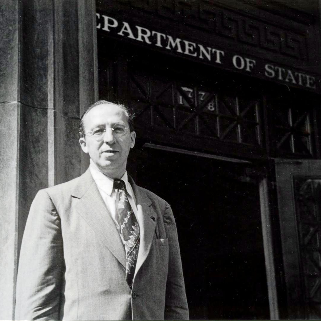 Copland wearing a suit and tie and standing in front of doorway with sign above it reading U.S. Department of State,