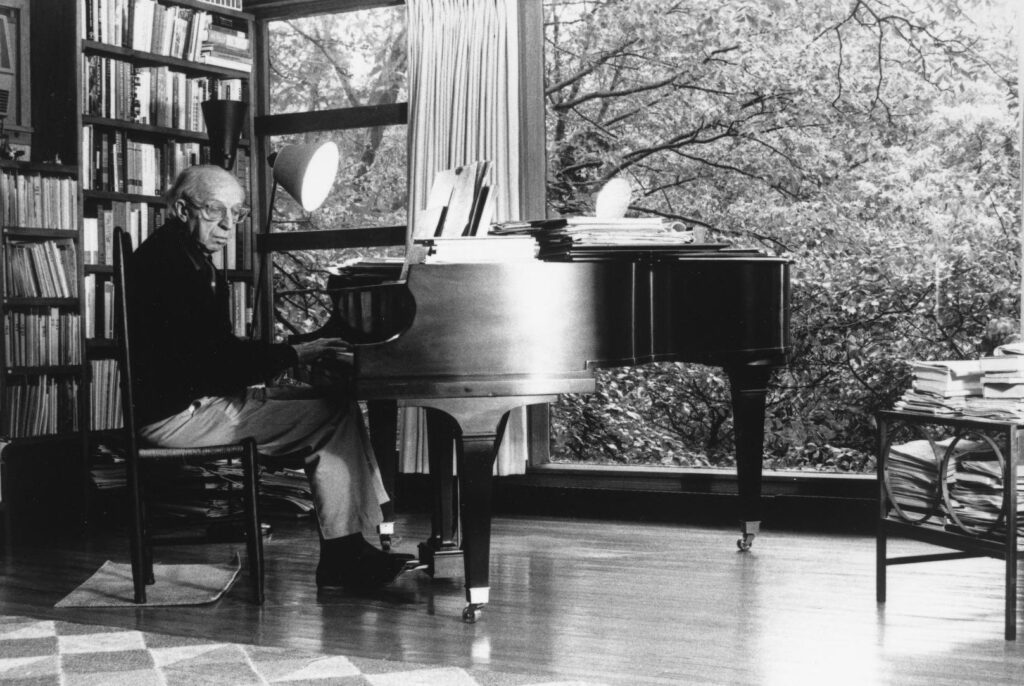 Aaron Copland sitting at grand piano and playing music in front of large window with bookshelves behind him.