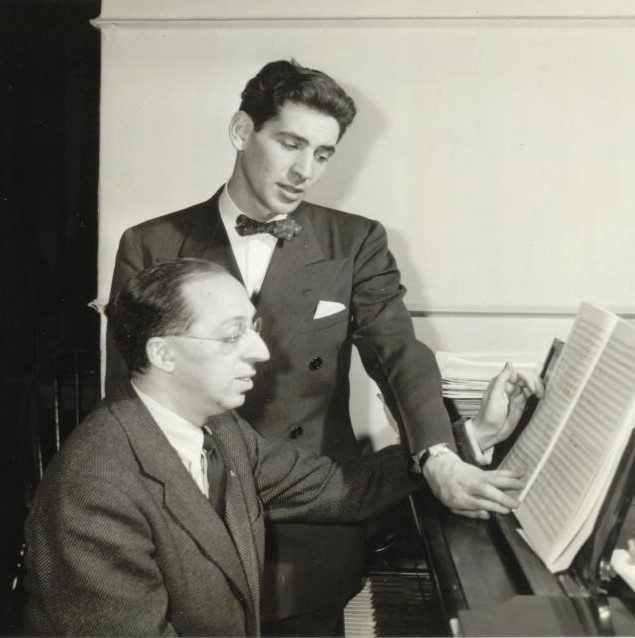 Aaron Copland sitting at piano wearing a suit, with composer Leonard Bernstein standing and reading sheet music over his left shoulder while he holds the page with his left hand.