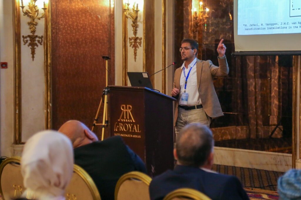 Alshami standing and speaking in a conference center in front of a podium pointing to his presentation on a screen above and behind him