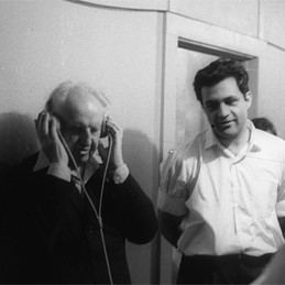 Copeland standing holding headphone speakers over his ears, listening to music with Amar Bose, music system inventor, looking on from his left.