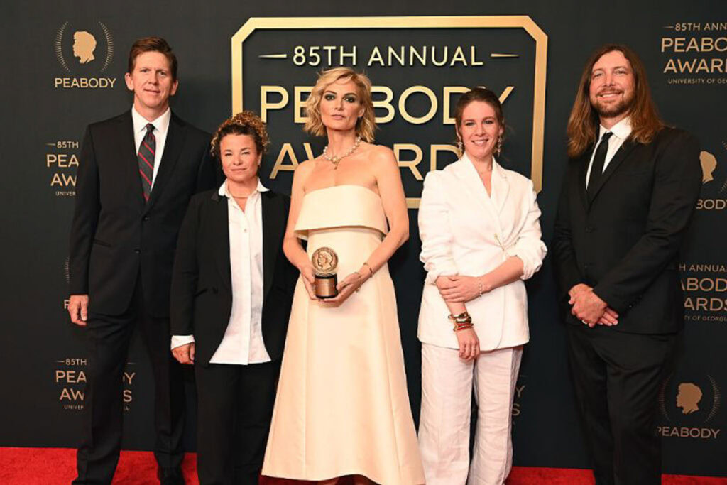Lucy Walker wearing an evening gown and holding her award, standing with four individuals on a red carpet at the 85th Annual Peabody Awards, with Peabody Awards logos and emblem on the dark backdrop behind them