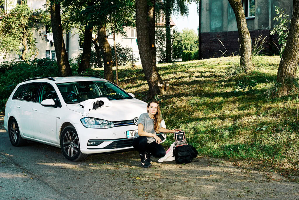 Marzena Abrahamik crouching next to a white station wagon on a sunny street with trees and houses in the background