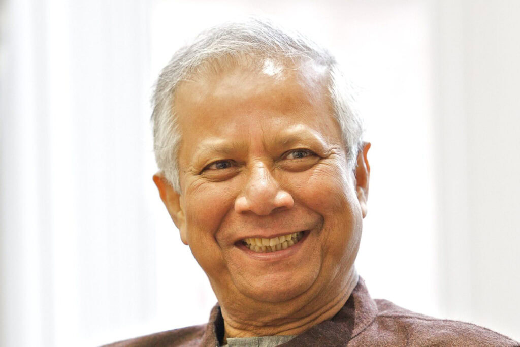 Nobel Peace Prize winner Muhammad Yunus with light gray hair and a brown suit jacket, smiling in front of a white background