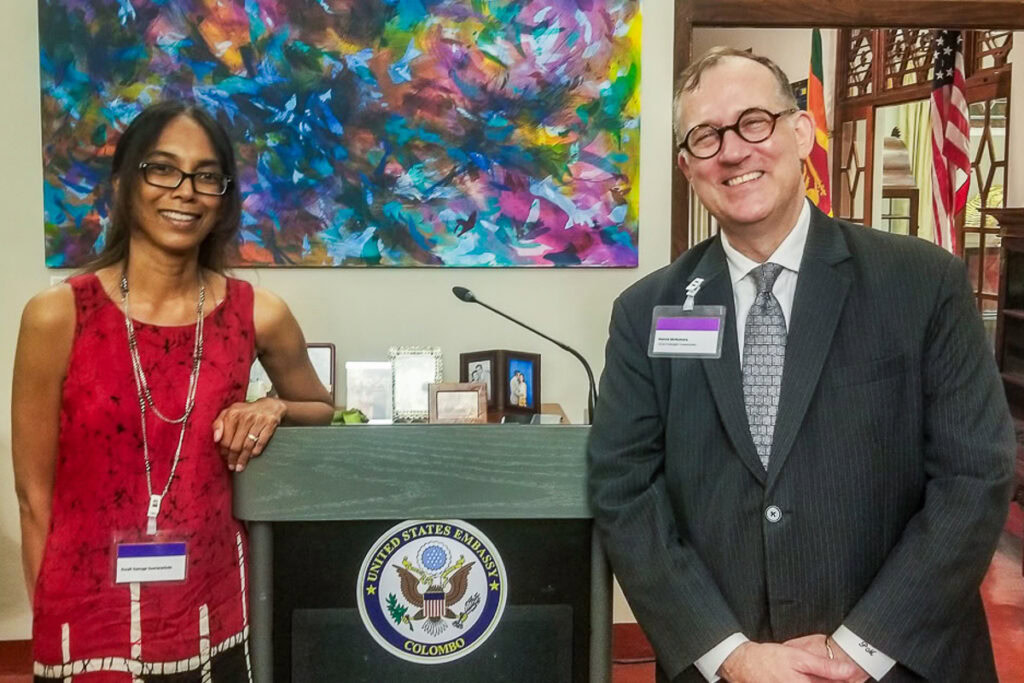 Kusali Gamage wearing a red sleeveless dress standing and smiling with a man in a gray suit in front of a podium with a State Department seal, in front of an abstract painting