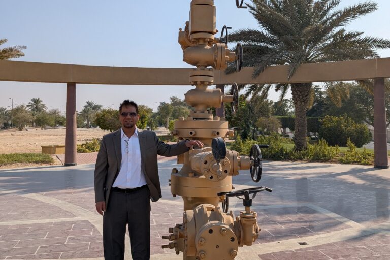 Ali Alshami standing next to a large industrial valve structure at a wellhead, outdoors in front of a date palm tree