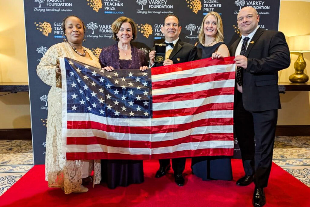 Five people holding an American flag on a red carpet in front of a backdrop with "Varkey Foundation" and "Global Teacher Prize" logos.