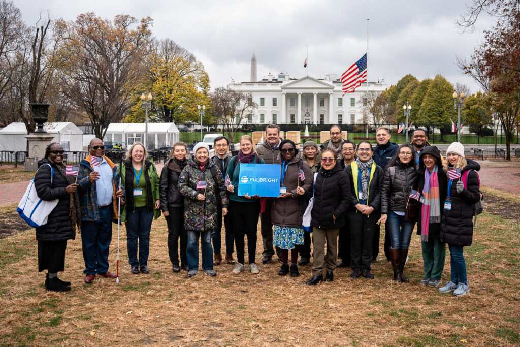 A diverse group of people stand smiling in front of the White House, some holding small American flags and a "Fulbright" sign, with a cloudy sky above.