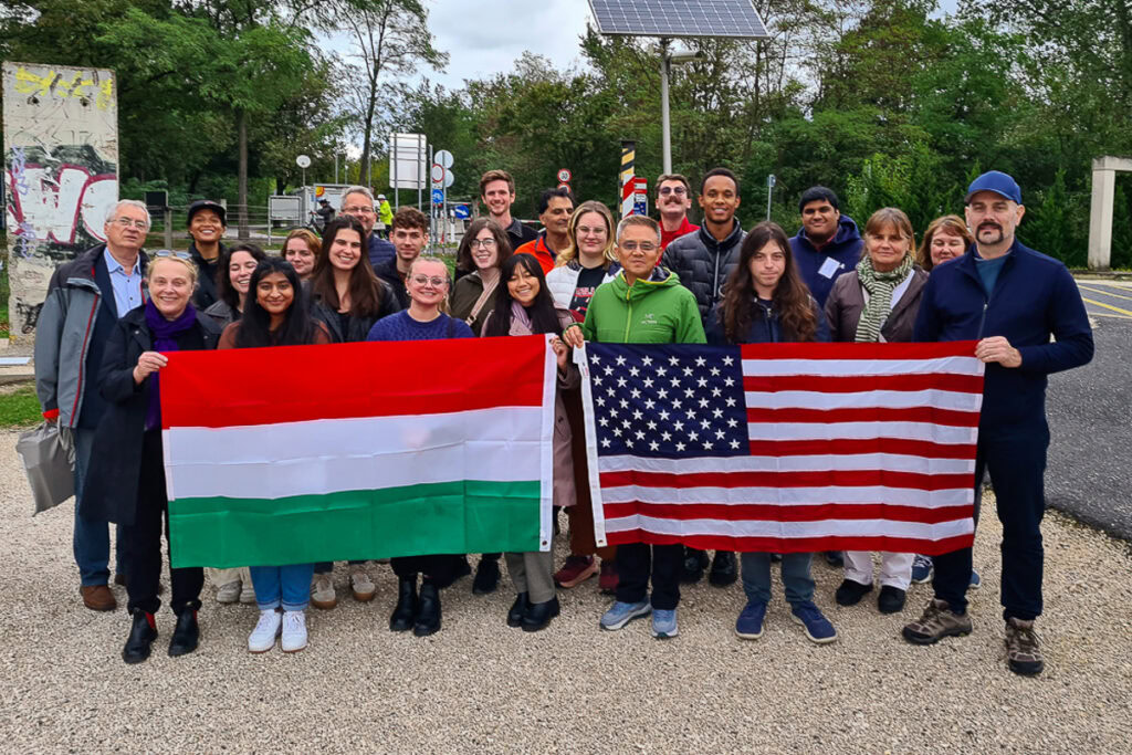 A group of Fulbright participants holding Hungarian and American flags stand on a gravel path in Hungary with trees behind them