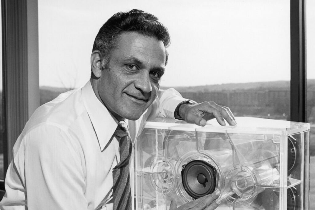 Amar Bose, wearing a dress shirt and tie, posing with a transparent speaker prototype revealing internal components against an office window with a cityscape backdrop