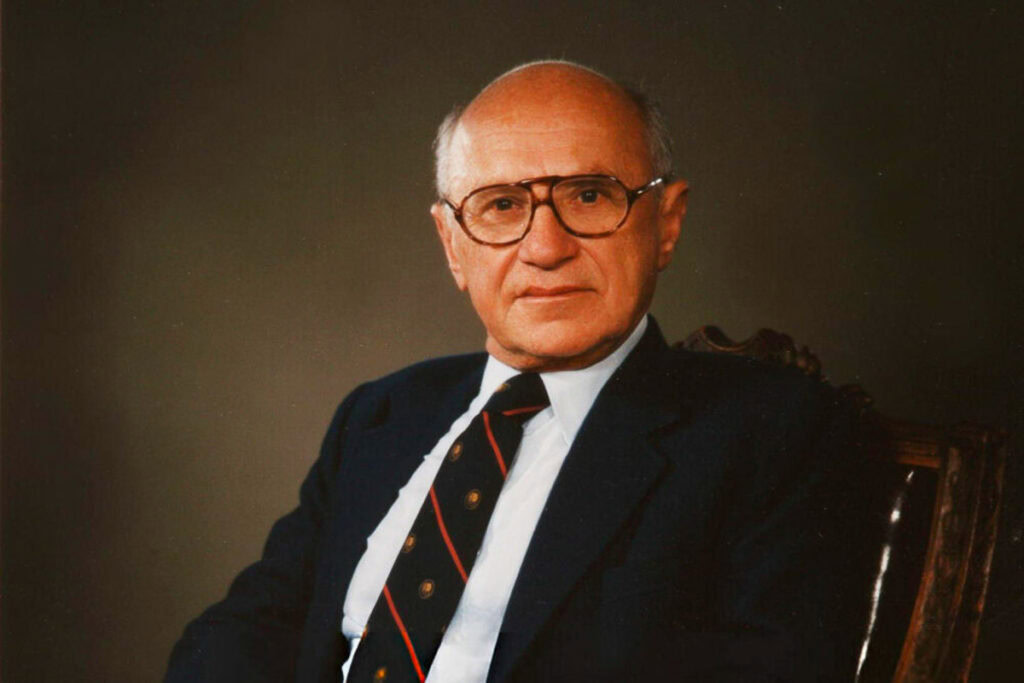 Milton Friedman wearing a dark suit and striped tie and glasses, seated in a large desk chair against a dark background