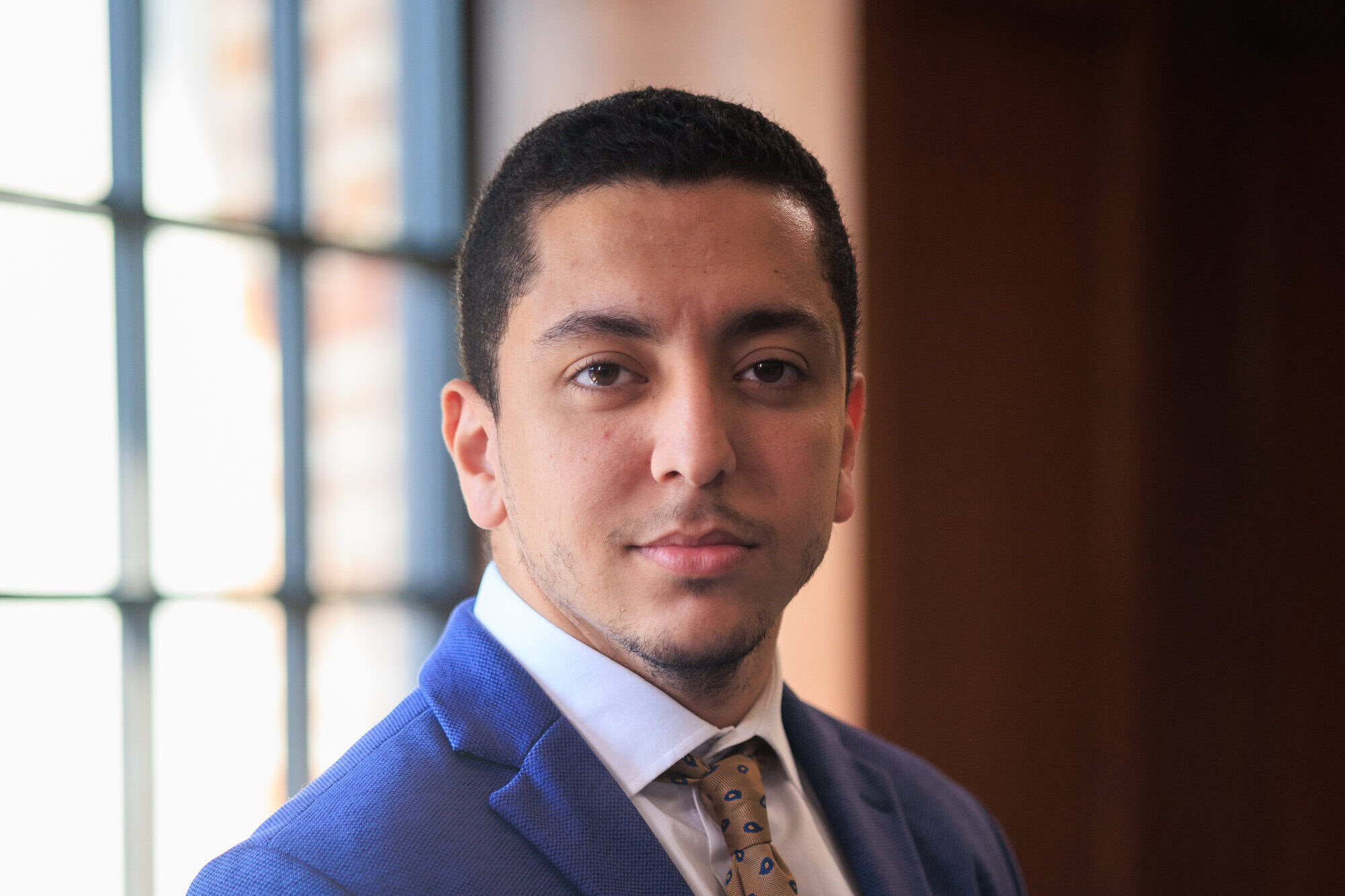 Khechai in a blue suit with a beige tie standing near a window.