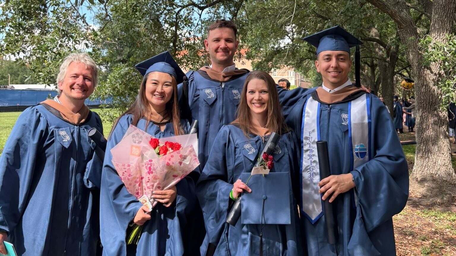 A group of five people wearing graduation gowns with two wearing graduation caps and two holding flowers standing outside in front of a tree